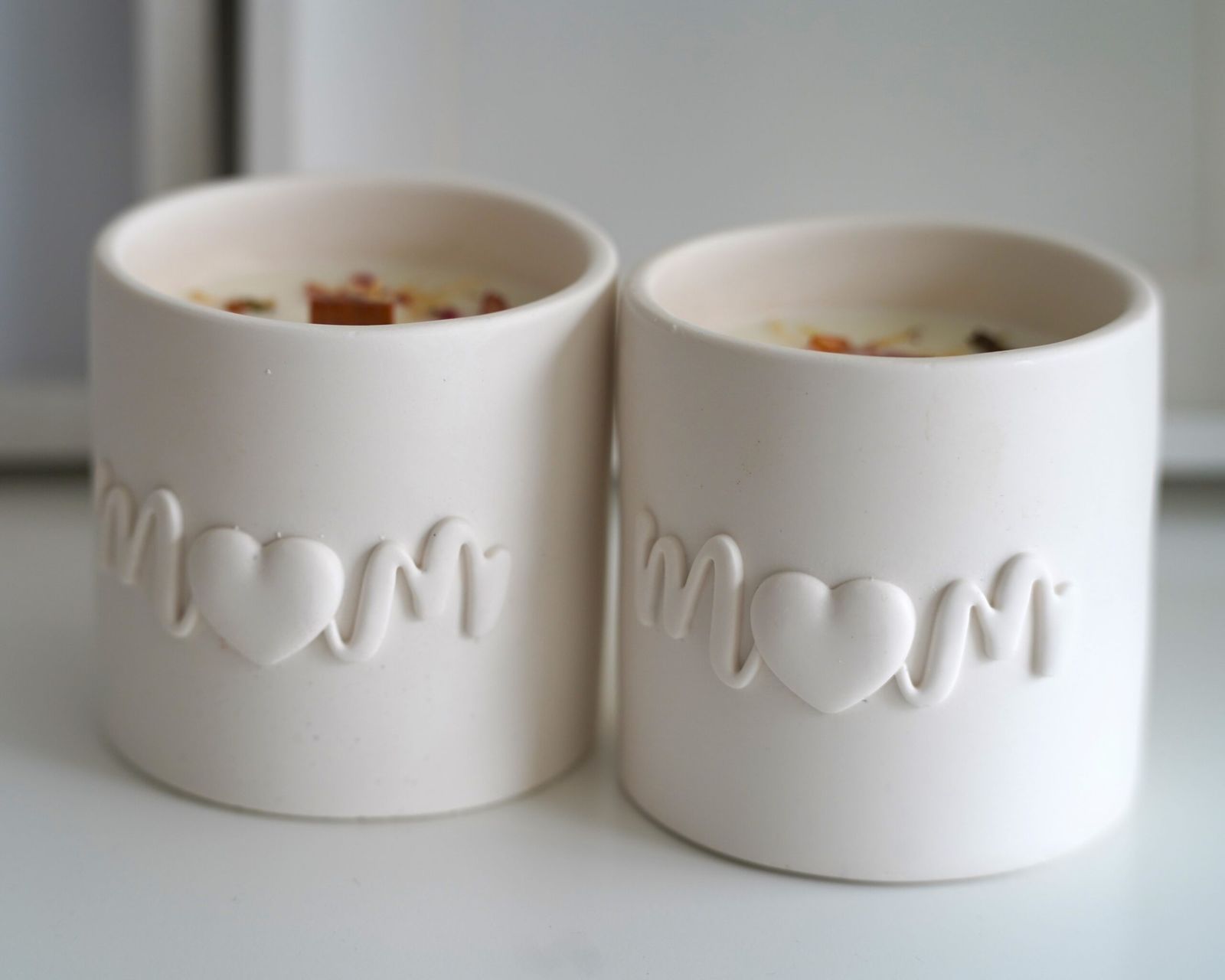 Velas artesanales de cera de soja en tarros de resina ecológica con la palabra "Mom" grabada. Vista frontal que deja entrever la cera y los pétalos de flores secas en su interior. Un aroma floral y afrutado en una edición limitada para el Día de la Madre.