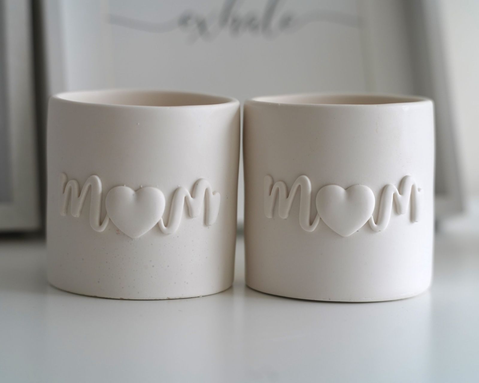 Dos velas artesanales en tarros de resina ecológica con la palabra "Mom" grabada en relieve. En su interior, una vela de cera de soja con un delicado aroma floral y afrutado. Edición limitada para el Día de la Madre.